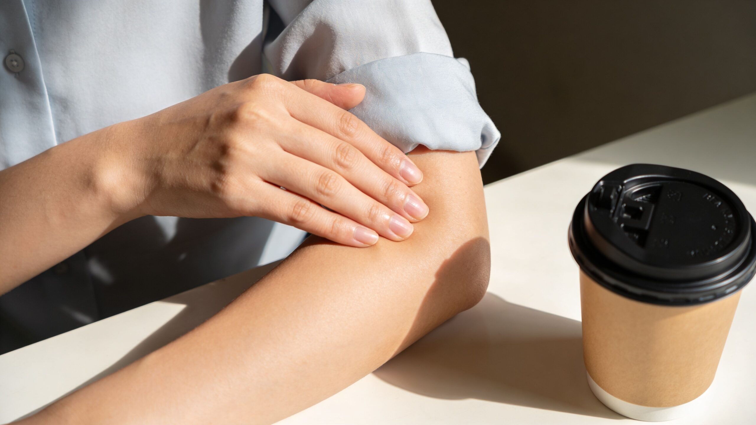 En kvinde masserer sin albue og arm, mens hun sidder ved et bord med en kop kaffe