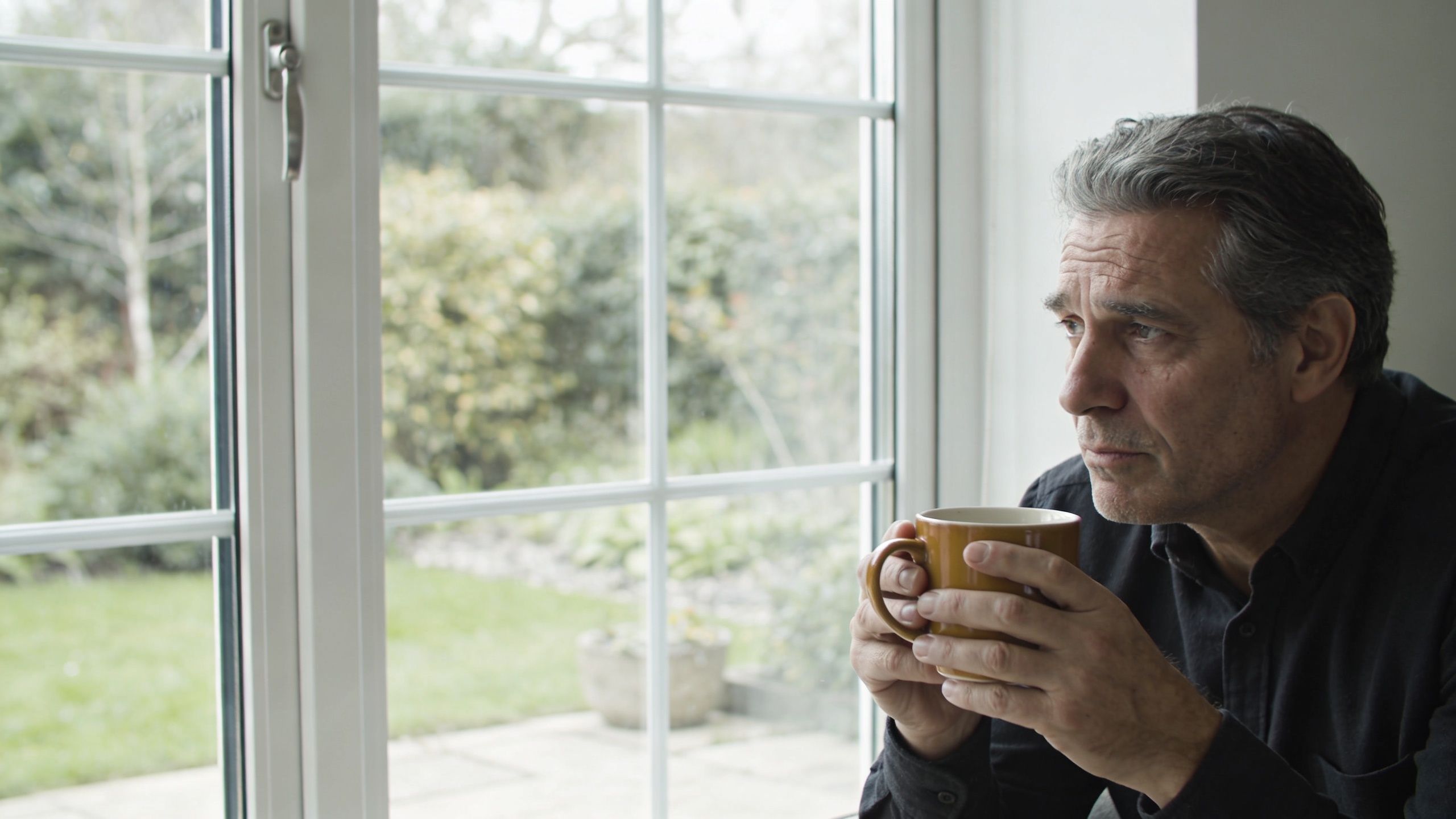 En ældre mand, der sidder eftertænksomt ved vinduet og holder en kop kaffe i sine hænder.