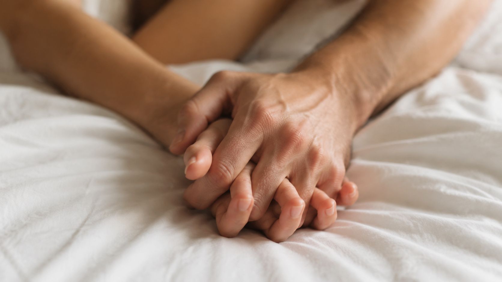 To personer holder hinanden i hænderne, mens de ligger i en seng med hvidt sengetøj.